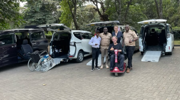 Bryce gives a thumbs up from his red powered mobility scooter, with the Accessible Travel Kenya team in front of four wheelchair-accessible vans with their ramps lowered